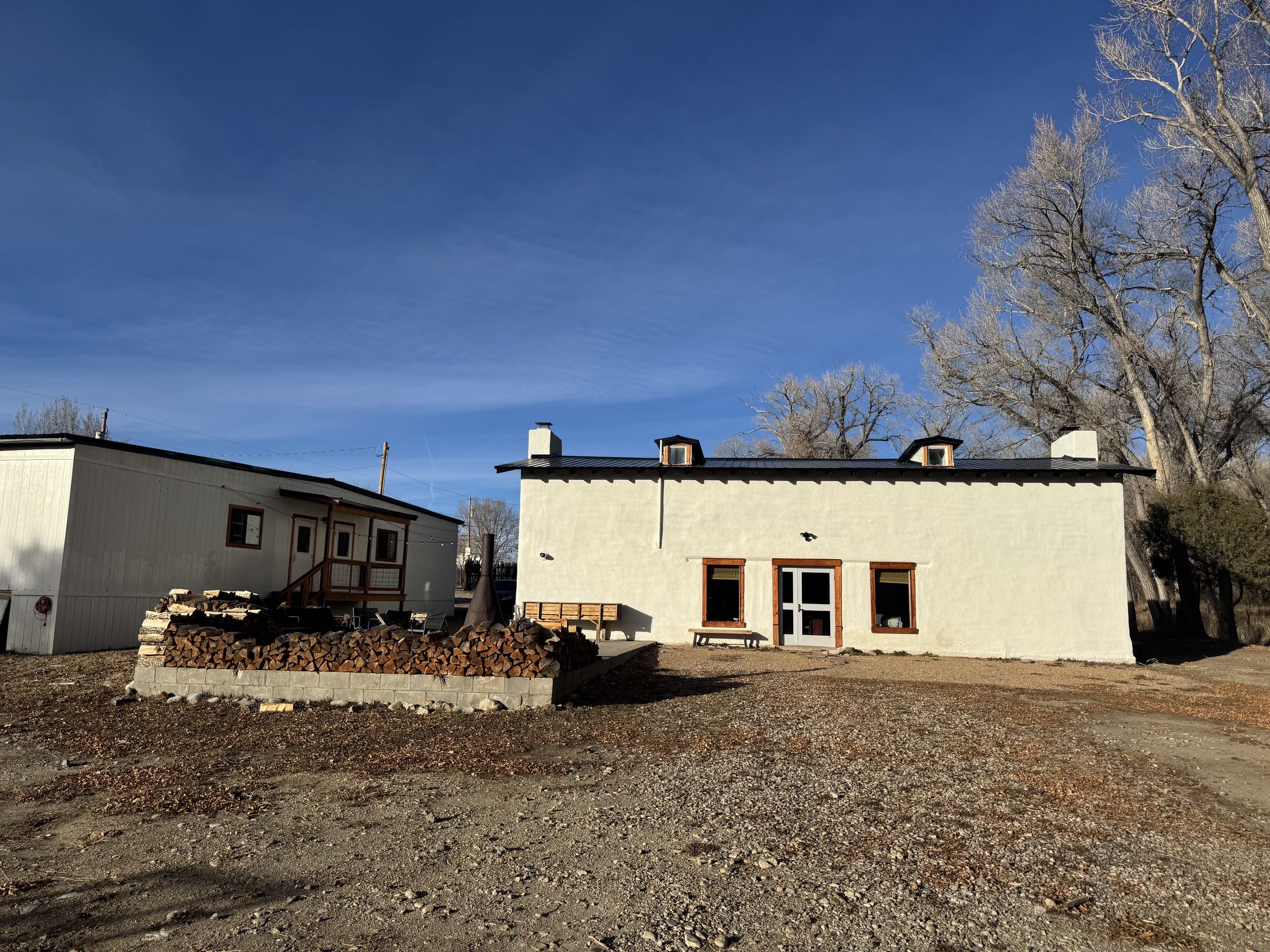 The white adobe home of River Community Church under a wide blue New Mexico sky, with stacked firewood and a bare cottonwood beside it in winter.