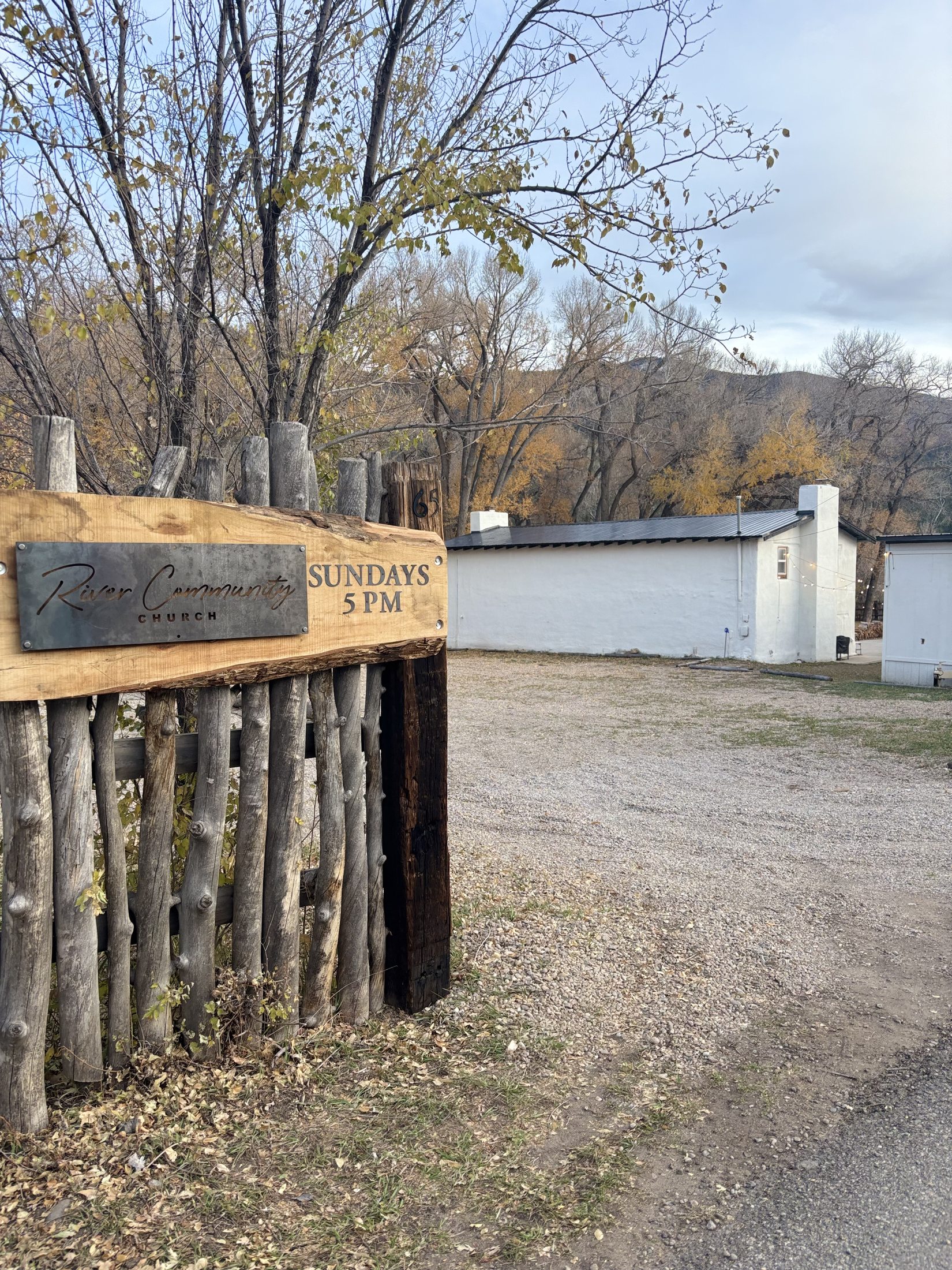 The approach to River Community Church from the road — a handcrafted wooden 'River Community Church, Sundays 5 PM' sign on a rustic latilla fence, with the white adobe building and the New Mexico mountains behind it.