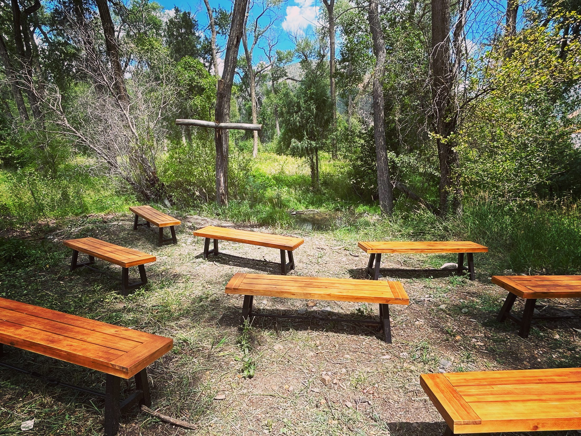 Handcrafted wooden benches arranged in a sunny clearing under the cottonwoods at River Community Church, facing a simple wooden cross.