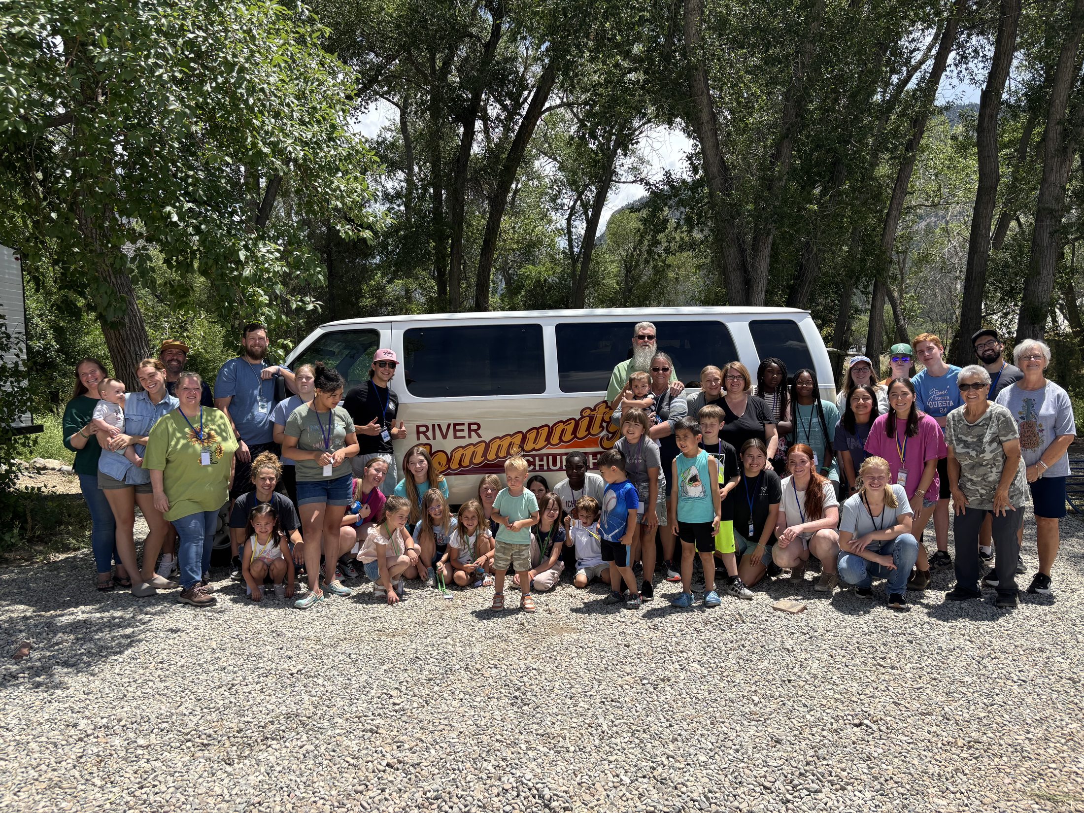 A large group of kids, students, and adults gathered in front of the River Community Church van on a summer day.