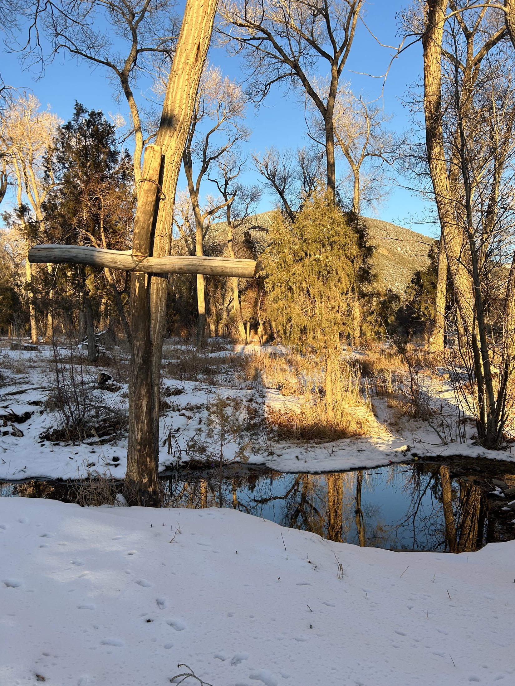 A simple wooden cross planted by a small creek on the church property, with snow on the banks and the New Mexico mountains rising in golden light behind it.