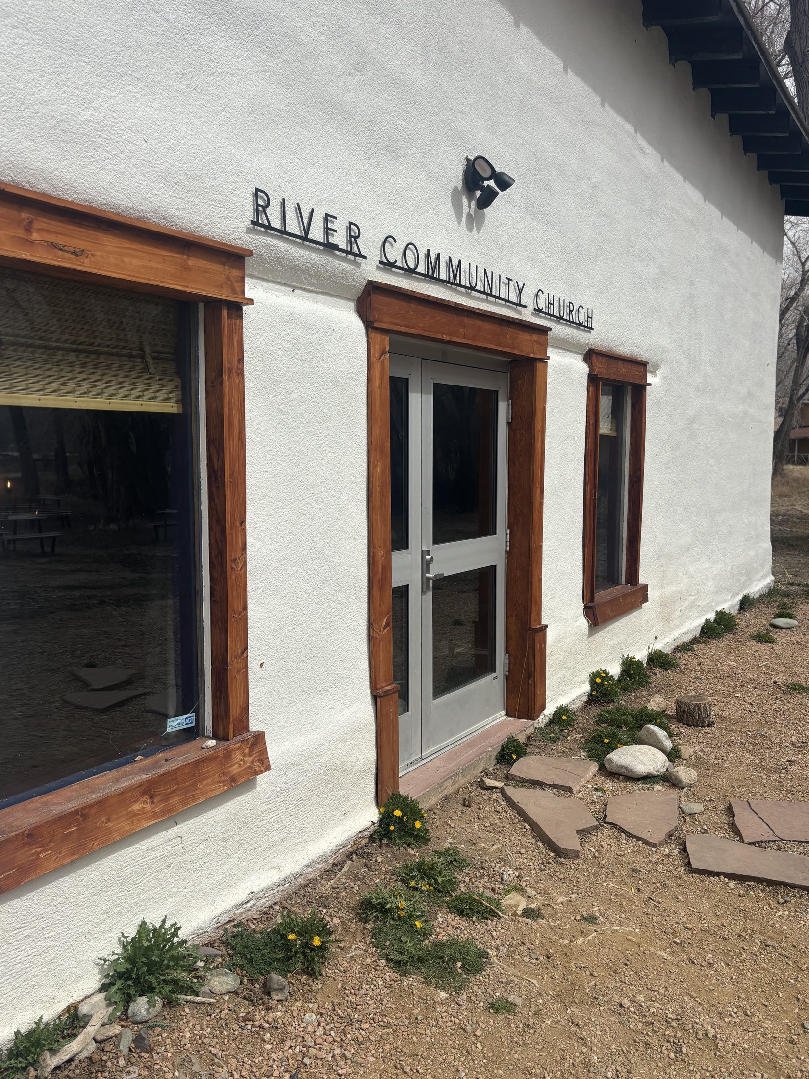 The front entrance of River Community Church — a white adobe building with wooden trim and the words RIVER COMMUNITY CHURCH set in dark metal letters above the door.
