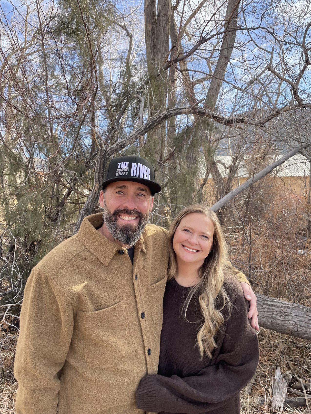 Pastor Jesse and Rachel Hardy standing together outdoors among the bare cottonwoods in Questa, New Mexico — Jesse in a brown jacket and a 'The River' cap, Rachel beside him.